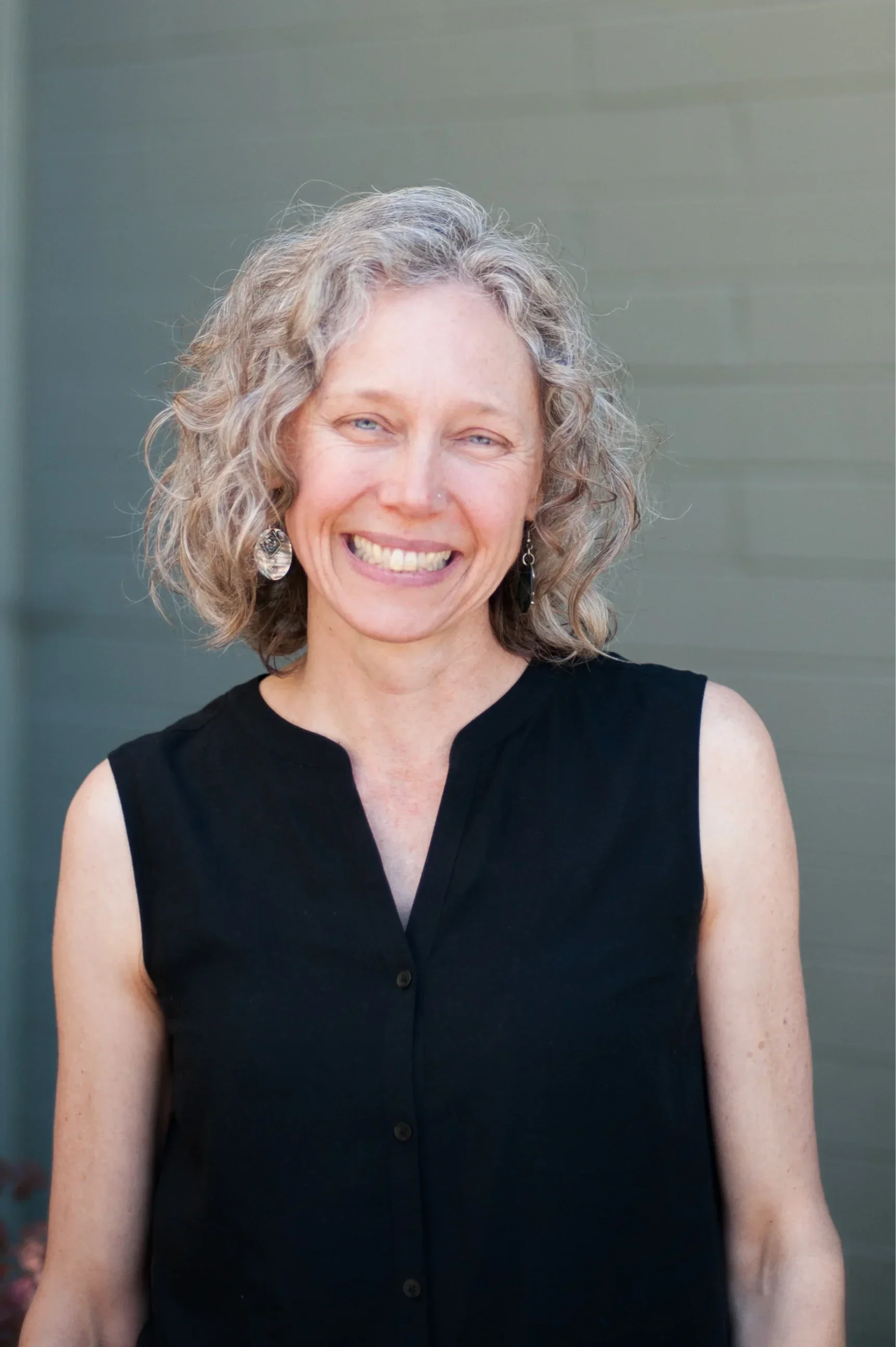 A woman with curly gray hair smiling, wearing a black sleeveless top and earrings, standing in front of a gray wall.