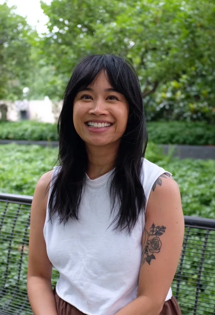 A woman with medium-length black hair and a tattoo of a rose and flowers on her upper arm, smiling outdoors with greenery in the background.