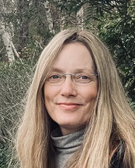 A woman with long blonde hair, glasses, and a gray turtleneck smiling outdoors in front of greenery and trees.