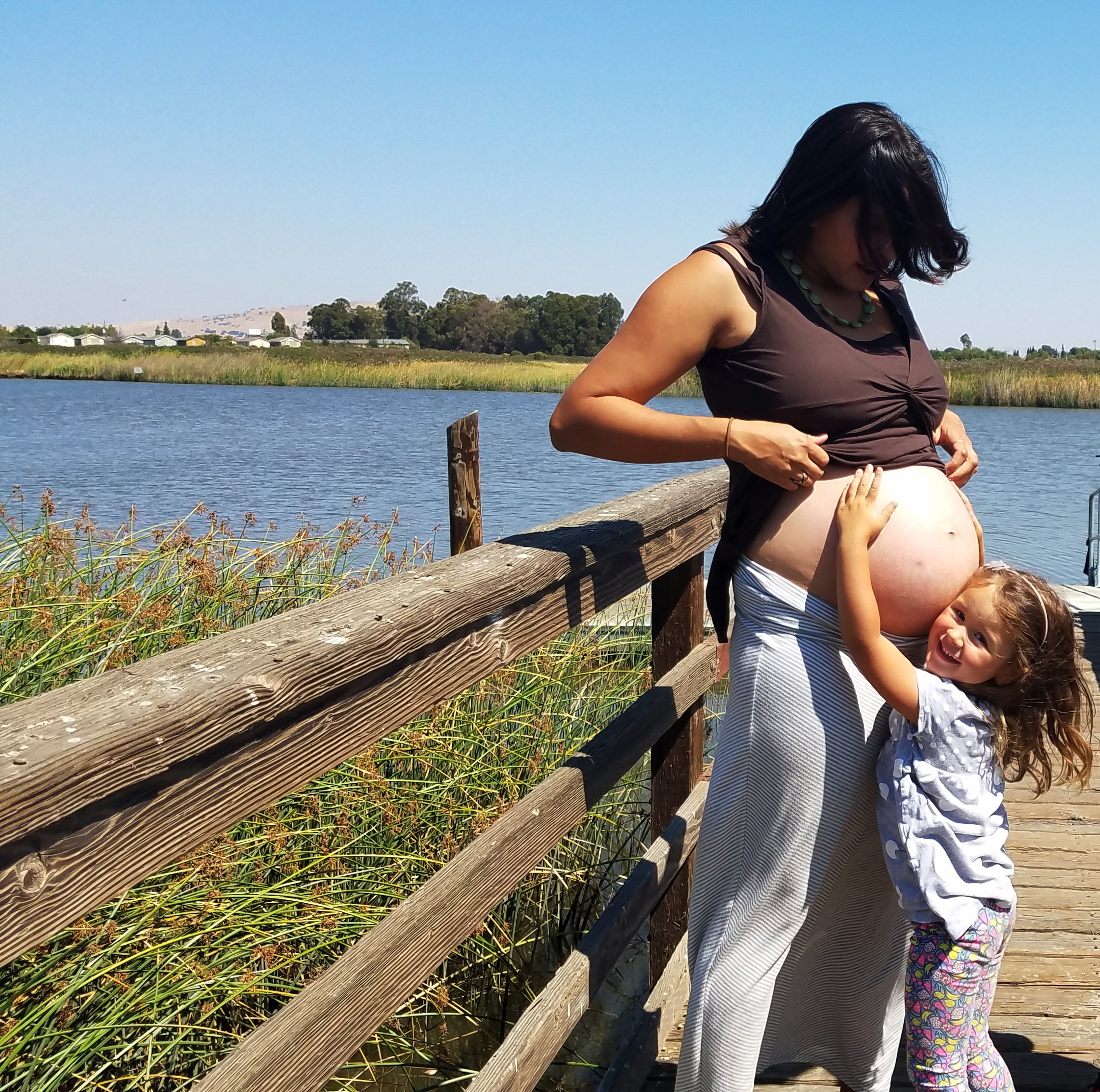 A pregnant woman stands on a wooden dock near a lake, wearing a brown top and striped skirt, with a young girl hugging her belly and smiling.