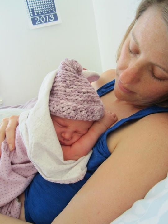 A woman holding a sleeping newborn baby on her chest, the baby wearing a pink knit hat and wrapped in a blanket.