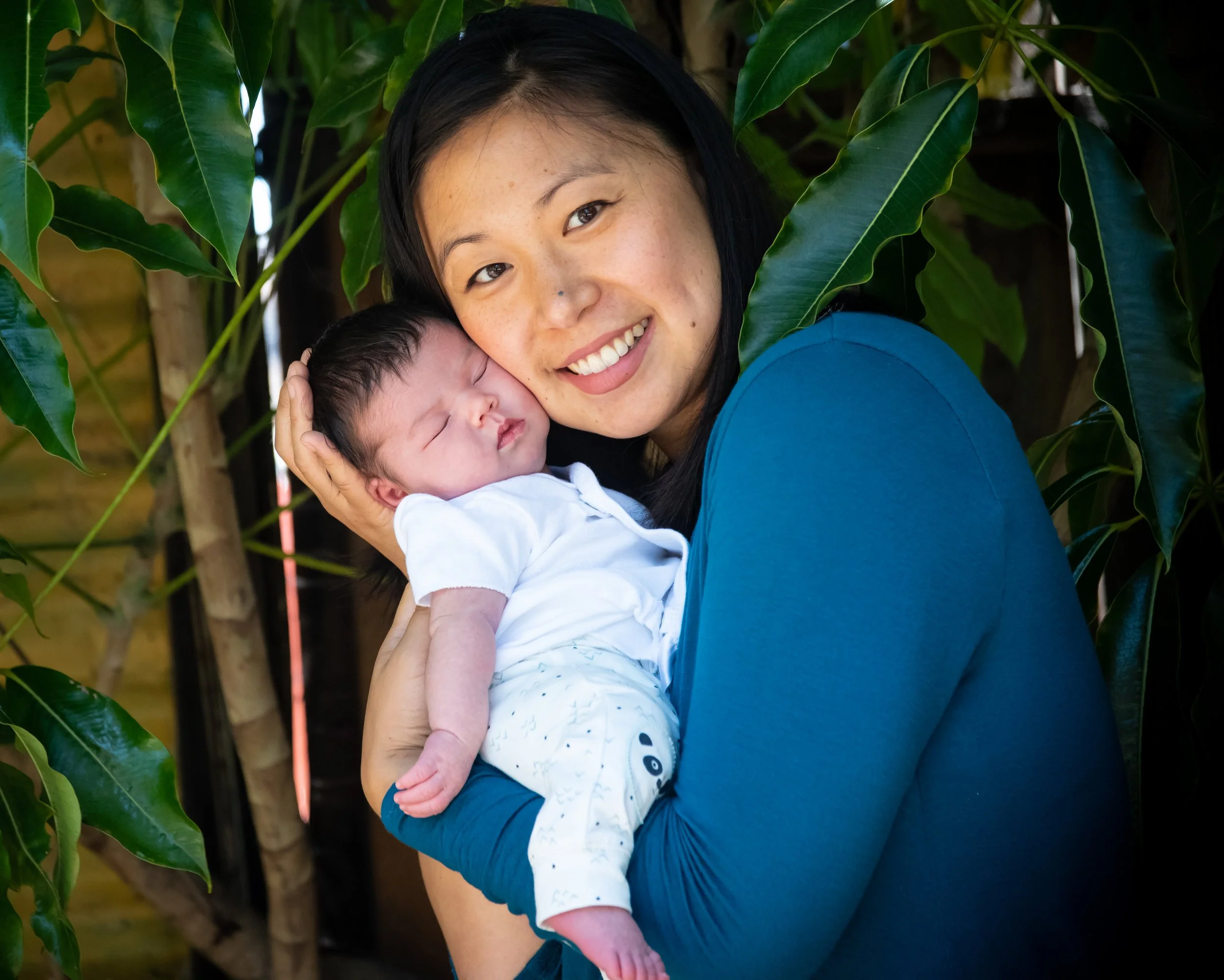 A woman holding a sleeping baby close to her face, smiling, with green leafy plants in the background.