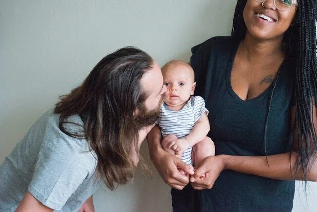 A man with long brown hair and a beard kisses a baby, held by a woman with long dark hair and glasses, in front of a plain wall.