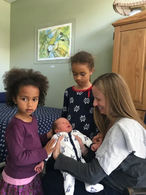 A happy family of three, including a man with glasses and a beard, a woman wearing a black hat and denim jacket, and a newborn baby with a white outfit, in a room with green walls and a colorful painting.