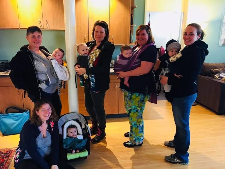 Group of six women and children standing in a kitchen and living room area, with some holding infants or toddlers and one sitting in a stroller, smiling at the camera.