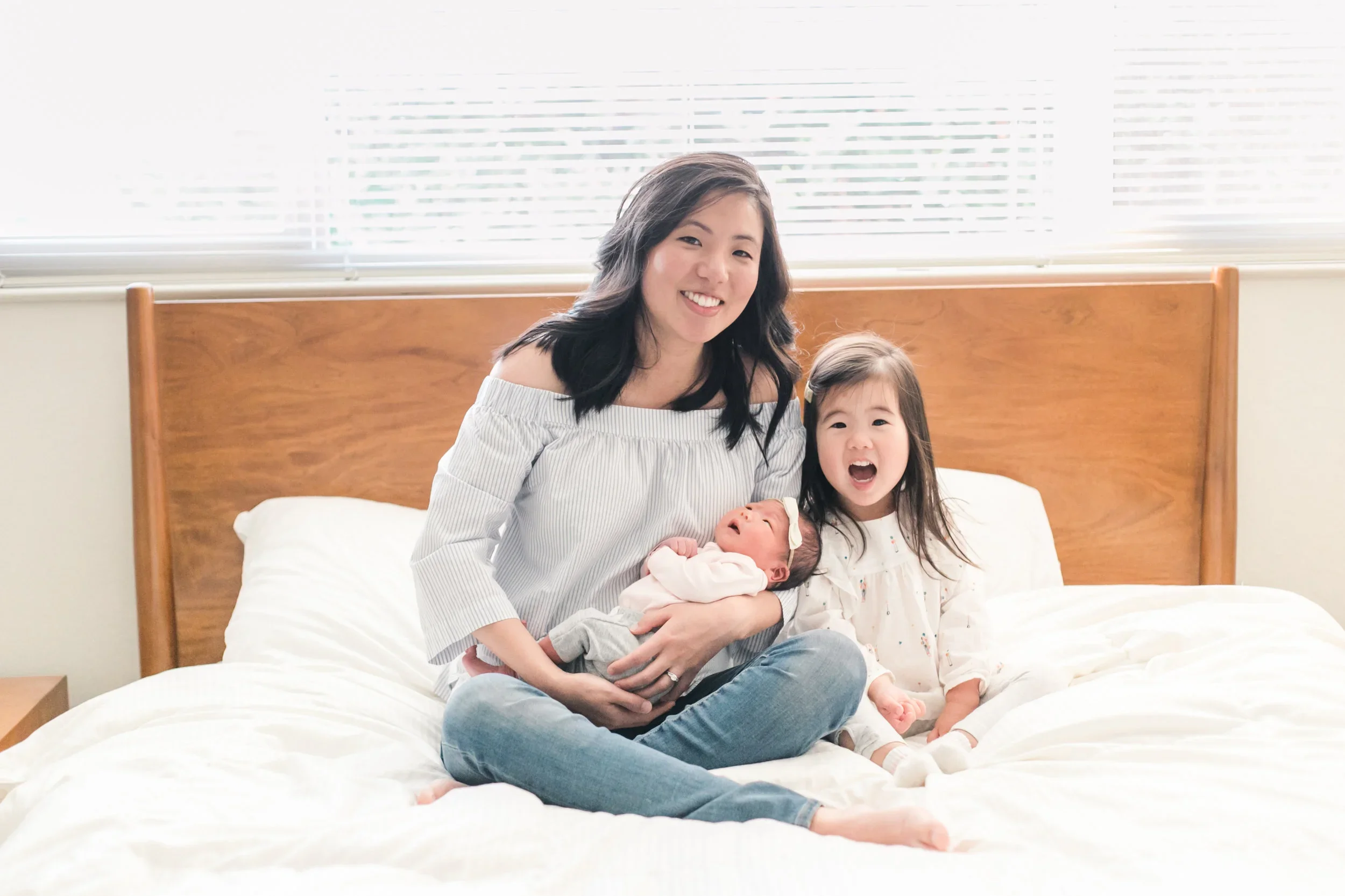 A woman, a young girl, and a newborn baby sitting on a bed in a bright bedroom.