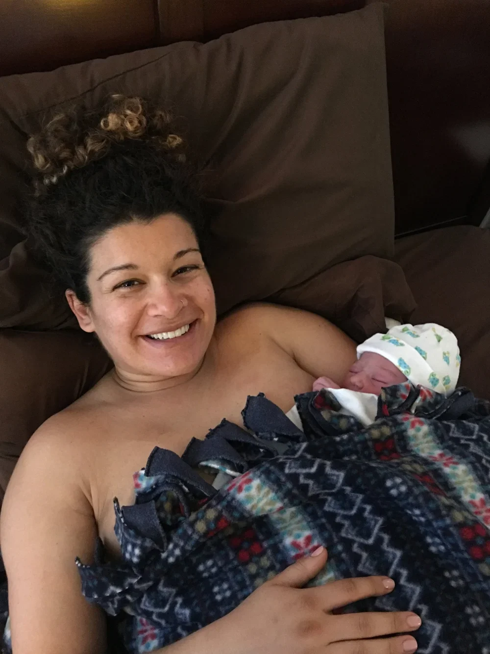 A woman lying in bed, smiling and breastfeeding a newborn baby wrapped in a blanket.