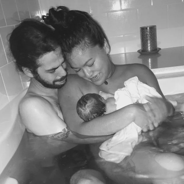A couple with a newborn in a bathtub, embracing and smiling, with tiled bathroom background and a candle in a holder.