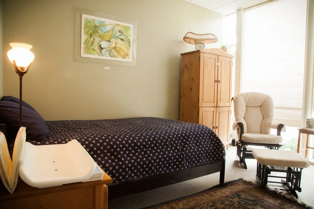 A bedroom with a bed covered in a black and white star patterned quilt, a wooden dresser, a cream-colored rocking chair and ottoman, a tall wooden cabinet, an abstract painting on the wall, a floor lamp, and sunlight coming through window blinds.