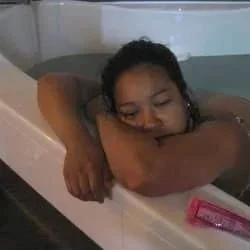 Woman relaxing in a bathtub, resting her head on her arm with a pink object nearby.
