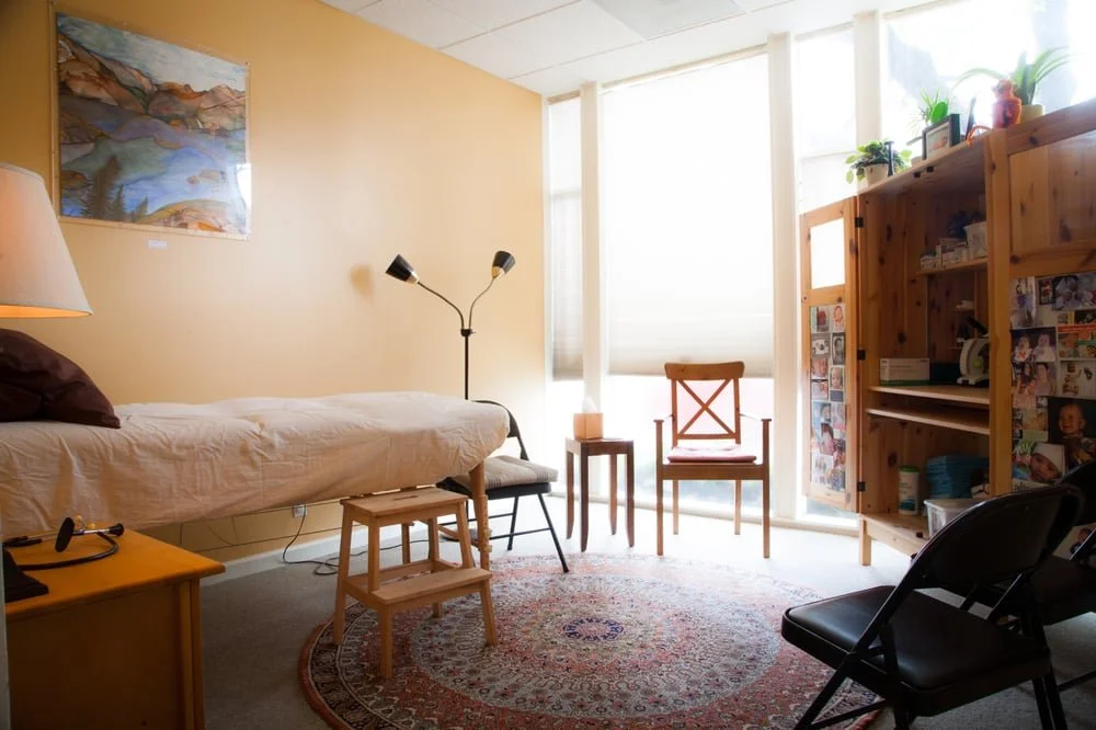 A sunny room with a bed, a wooden chair, a small table, a bookshelf filled with photos and items, a black chair, and a colorful rug, with large windows covered by white blinds.