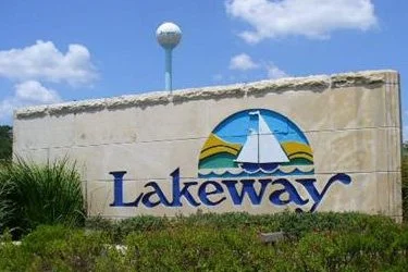 A large stone sign with the word 'Lakeway' and a sailboat logo, located in front of a grassy area under a blue sky.