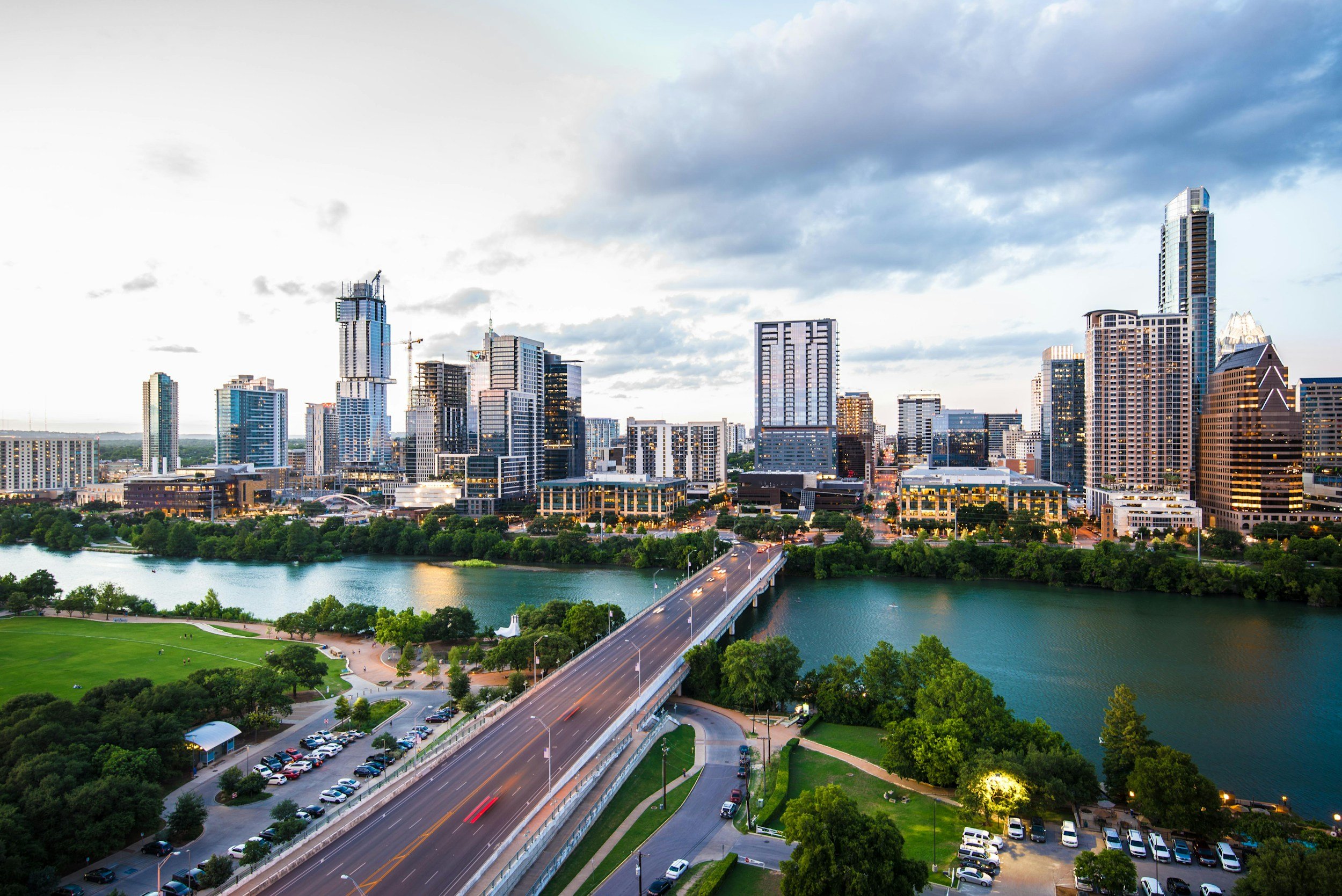 A city skyline with tall skyscrapers, a bridge over a river, and a park with green trees and a parking lot, during dusk.