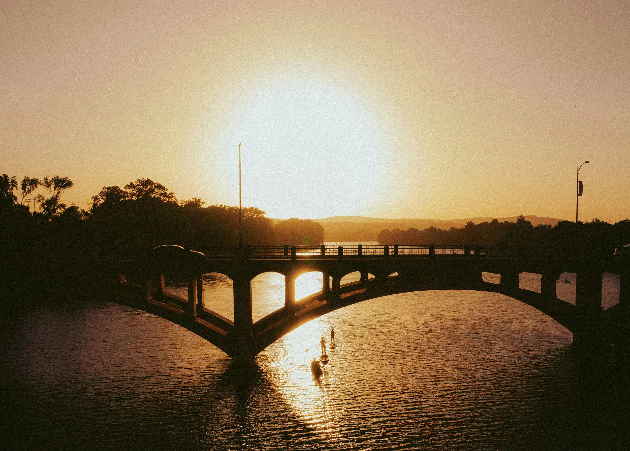 Sunset over a bridge crossing a calm river, with paddle boats and silhouettes of trees in the background.
