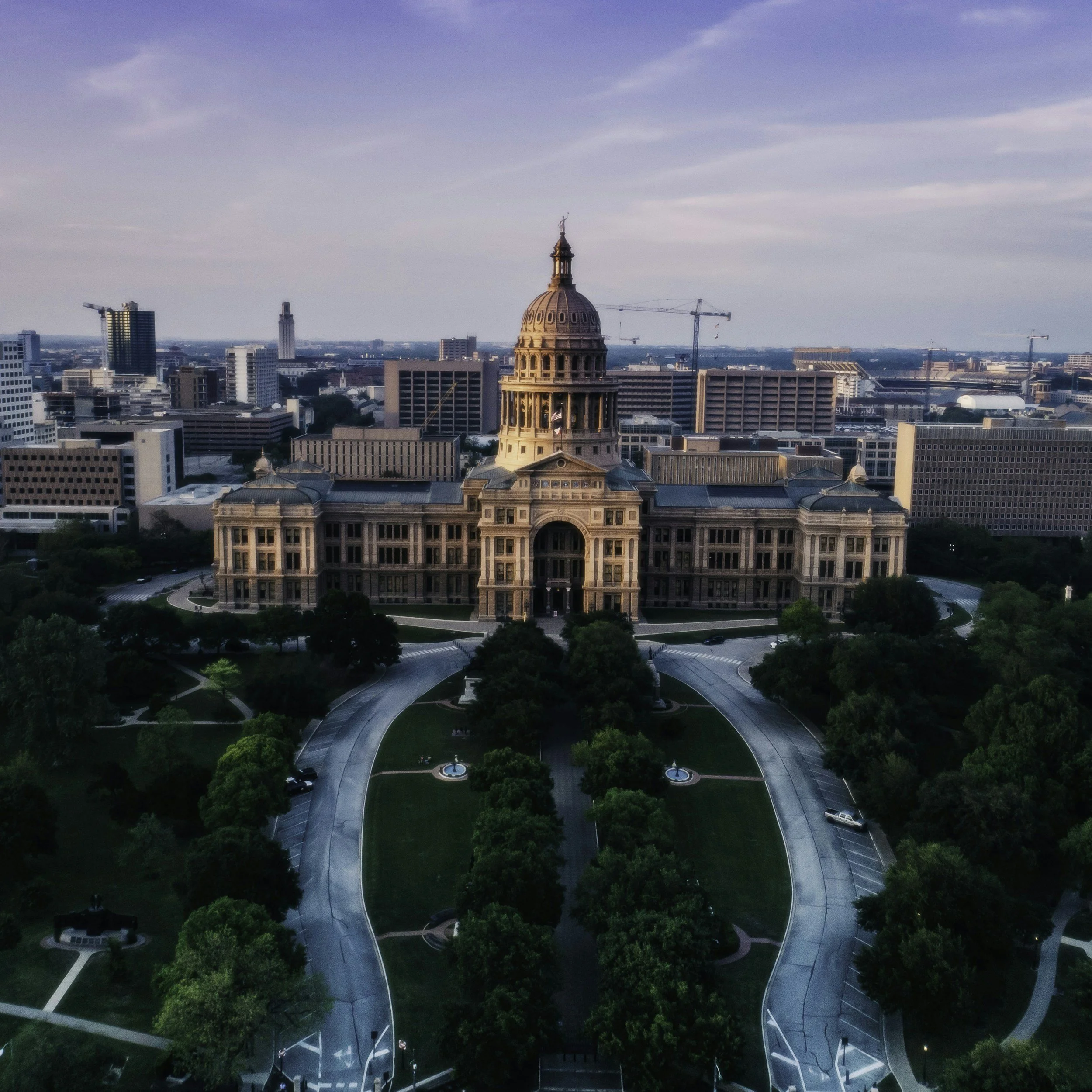 Aerial view of a large historic government building with a dome, surrounded by a park with trees and walkways, in a cityscape with modern buildings in the background.