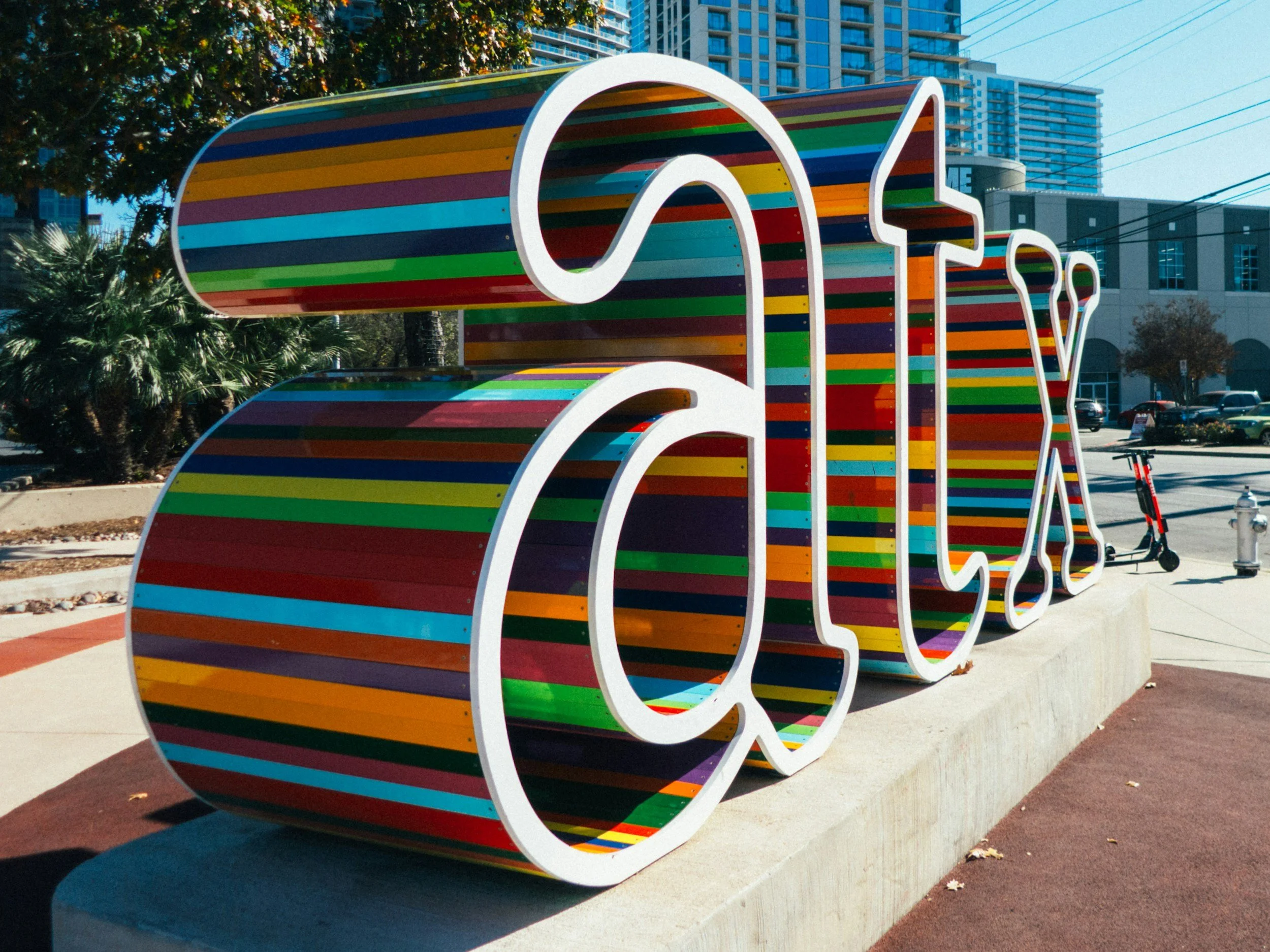 Colorful striped sculpture of the word 'LOVE' with large white outlines, situated outdoors in an urban area with trees, buildings, and electric wires in the background.