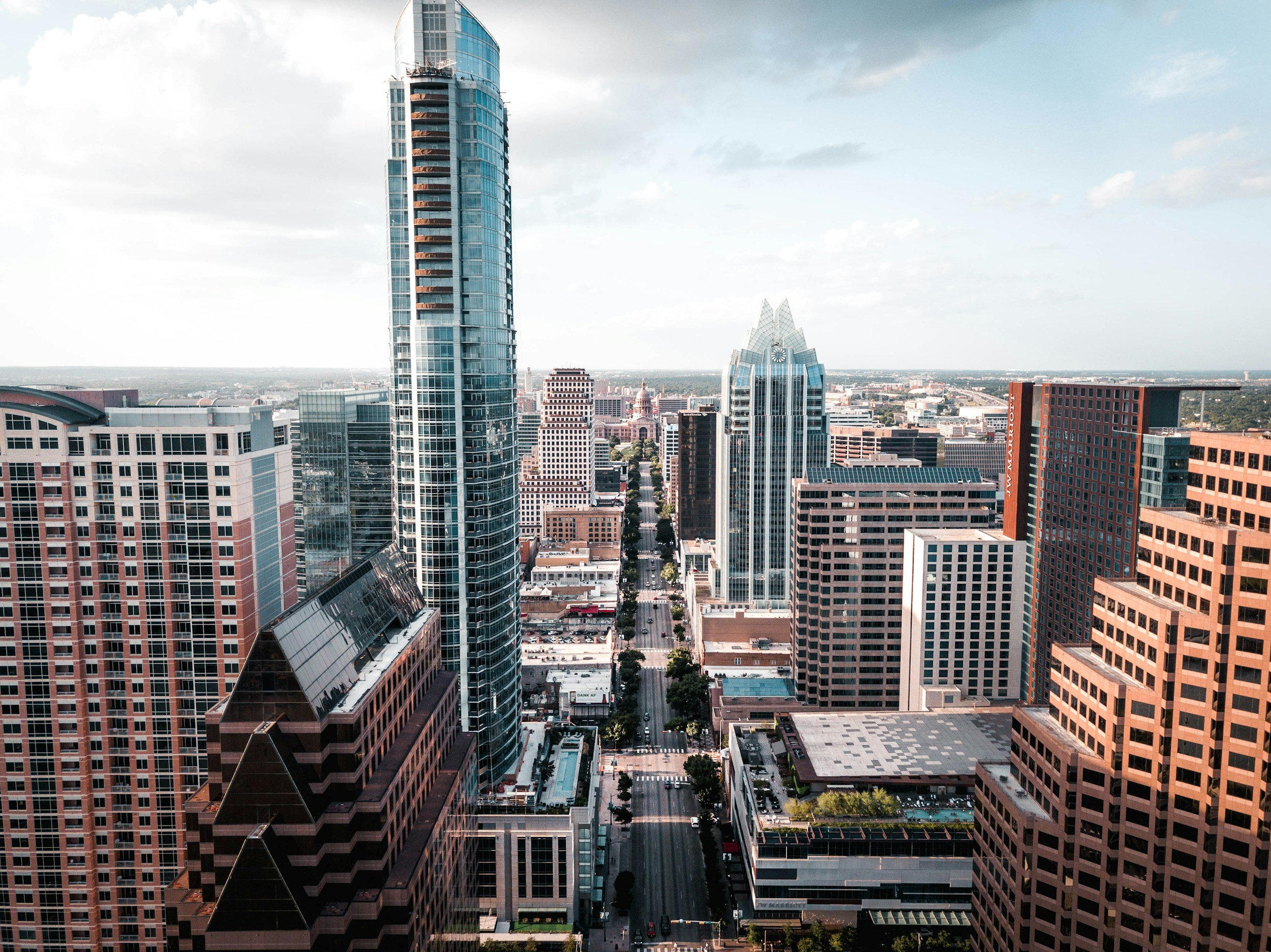 A city skyline with tall modern skyscrapers and office buildings, viewed from a high vantage point. Streets and trees are visible below.