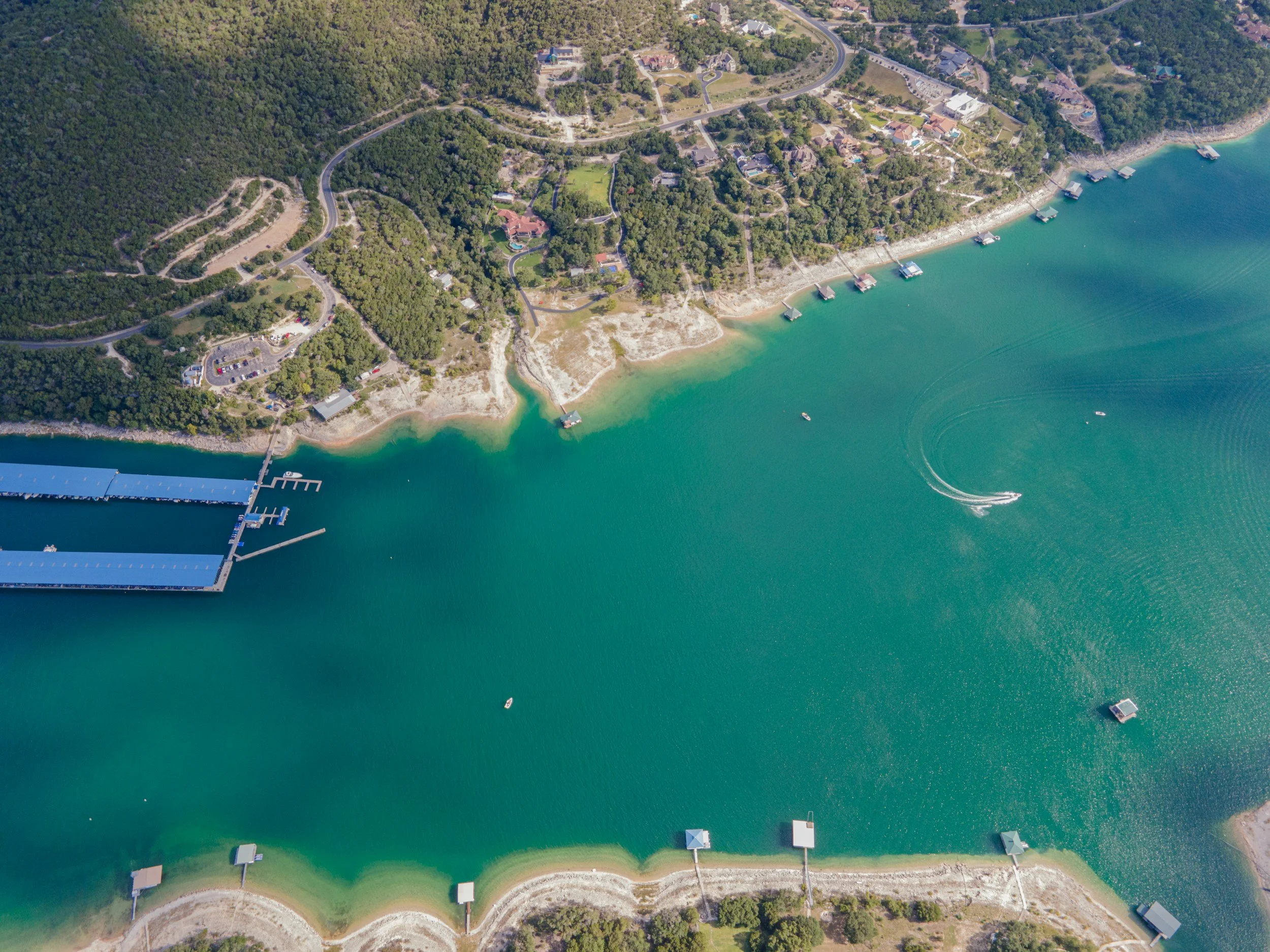 Aerial view of a marina with docked boats, a man-made lake with boats, and surrounding green forested hills with residential buildings and roads.