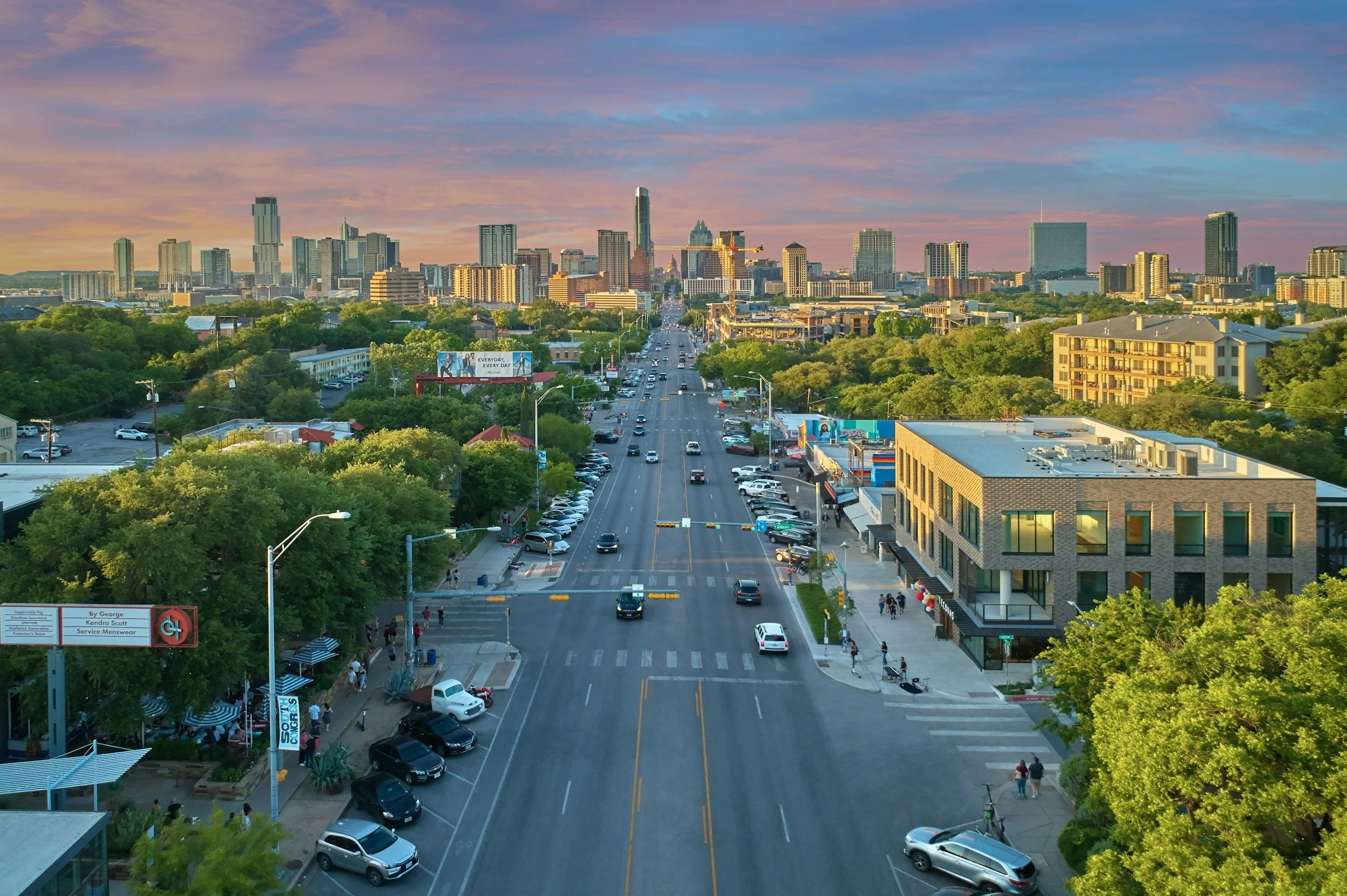 A city street view with cars driving, parked on the sides, trees lining the sidewalks, and a skyline of tall buildings in the background at sunset.