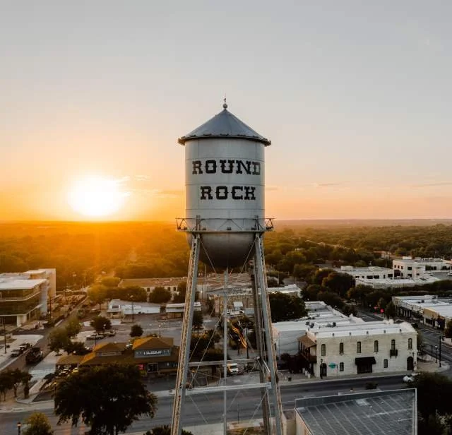A tall water tower with the words 'ROUND ROCK' painted on it, standing over a small town during sunset.