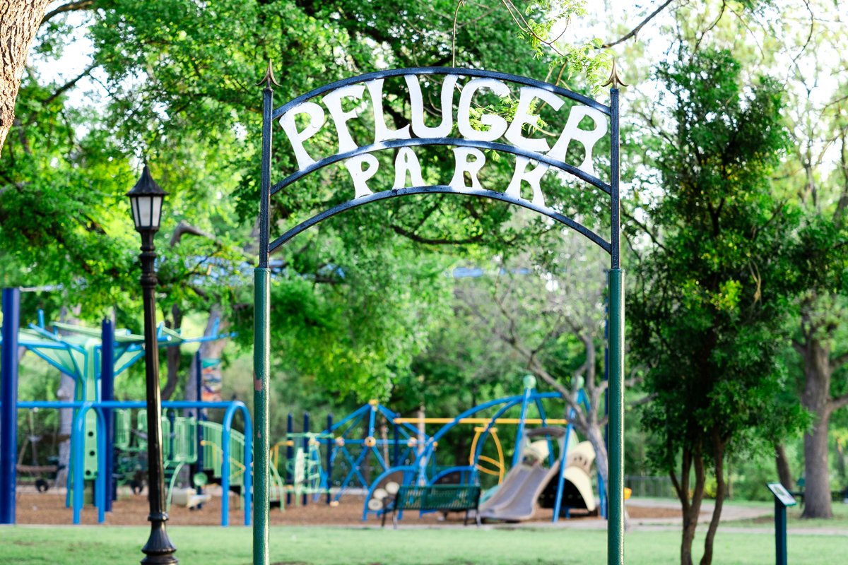 Sign at Puppeter Park with playground equipment, trees, and a lamp post in the background.