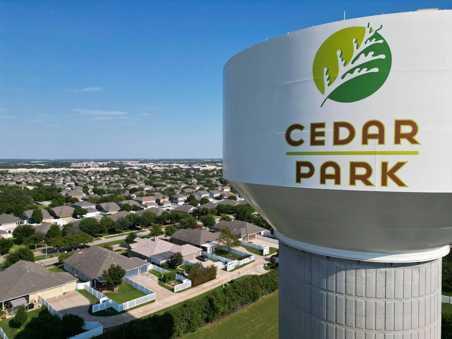 A large white water tower with the logo and name of Cedar Park in green and red, overlooking a neighborhood with single-family homes and trees.