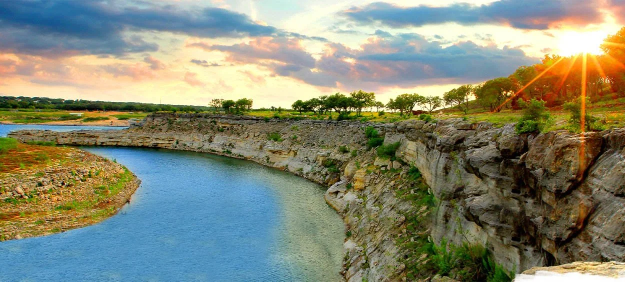 Sunset over a winding river flowing past rocky cliffs with trees on top, under a partly cloudy sky.
