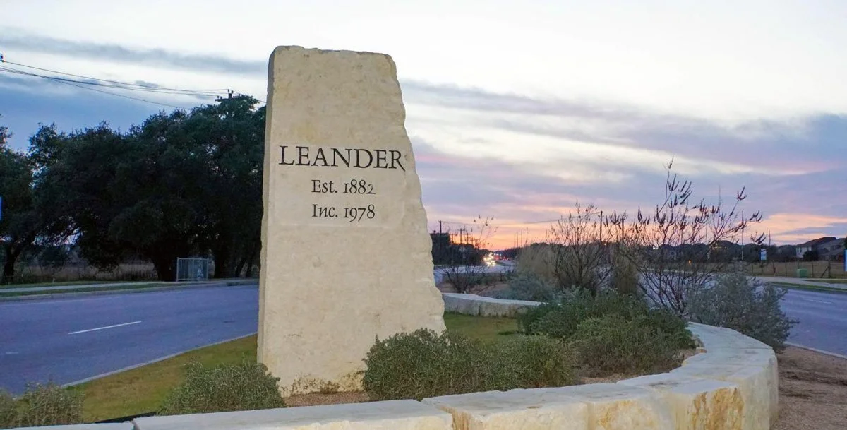 A large stone sign reads 'Leander, Est. 1882, Inc. 1978' and is situated on a landscaped area next to a curved road with trees and houses in the distance during sunset or sunrise.