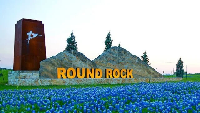 Sign that reads 'Round Rock' with rocks and flowers in the foreground, and a tall rust-colored monument with a jet plane sculpture on top in the background.