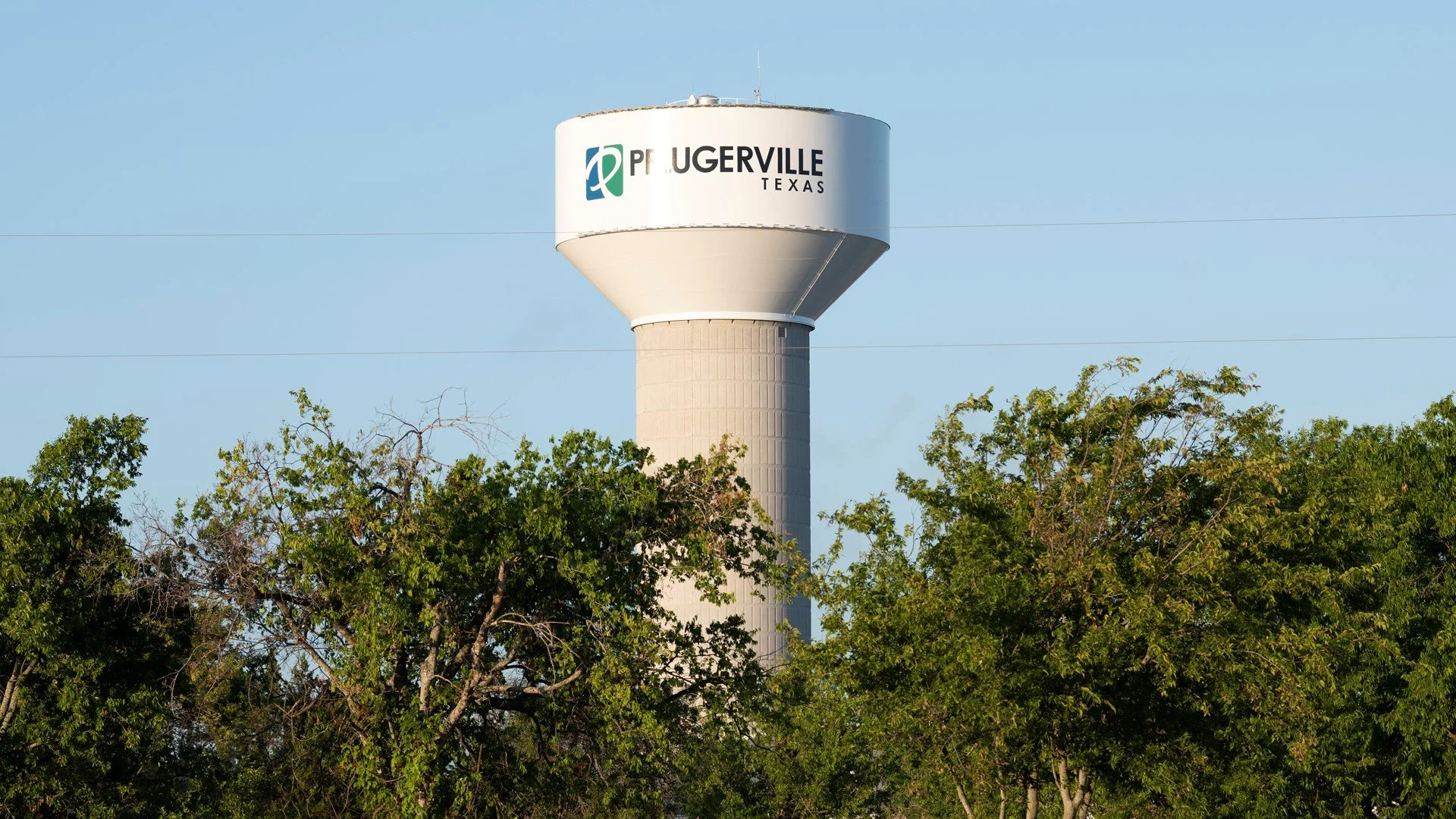 Water tower with a white cylindrical structure and a rounded top, with the words 'Peggyville Texas' on it, surrounded by green trees under a clear blue sky.