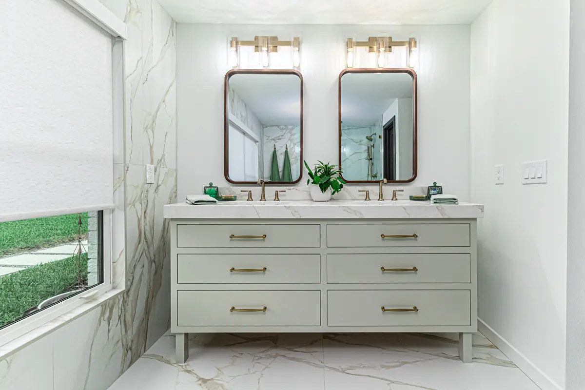 A modern bathroom featuring a double vanity with a white marble countertop, two rectangular mirrors with wooden frames, wall-mounted light fixtures, a potted plant between the mirrors, and a window with a view of green grass outside.