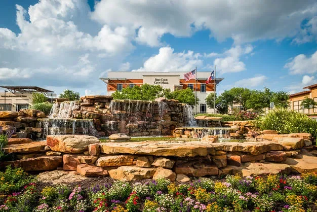 A landscaped outdoor area with a tiered rock fountain, colorful flowers in the foreground, and a building with a sign and American flags in the background.