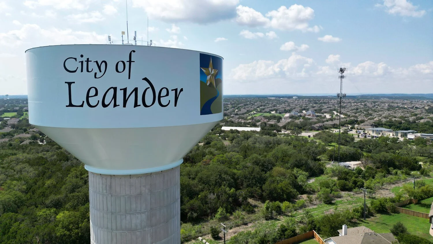 Elevated water tower with the logo and name of the City of Leander, Texas, in a suburban area with trees, houses, and a distant skyline.