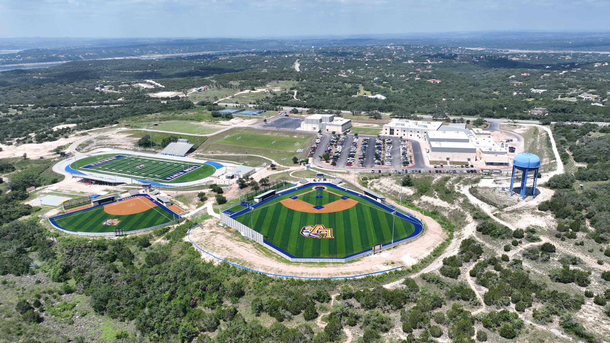 Aerial view of a sports complex with baseball and soccer fields, parking lots, and a water tower in a green landscape.