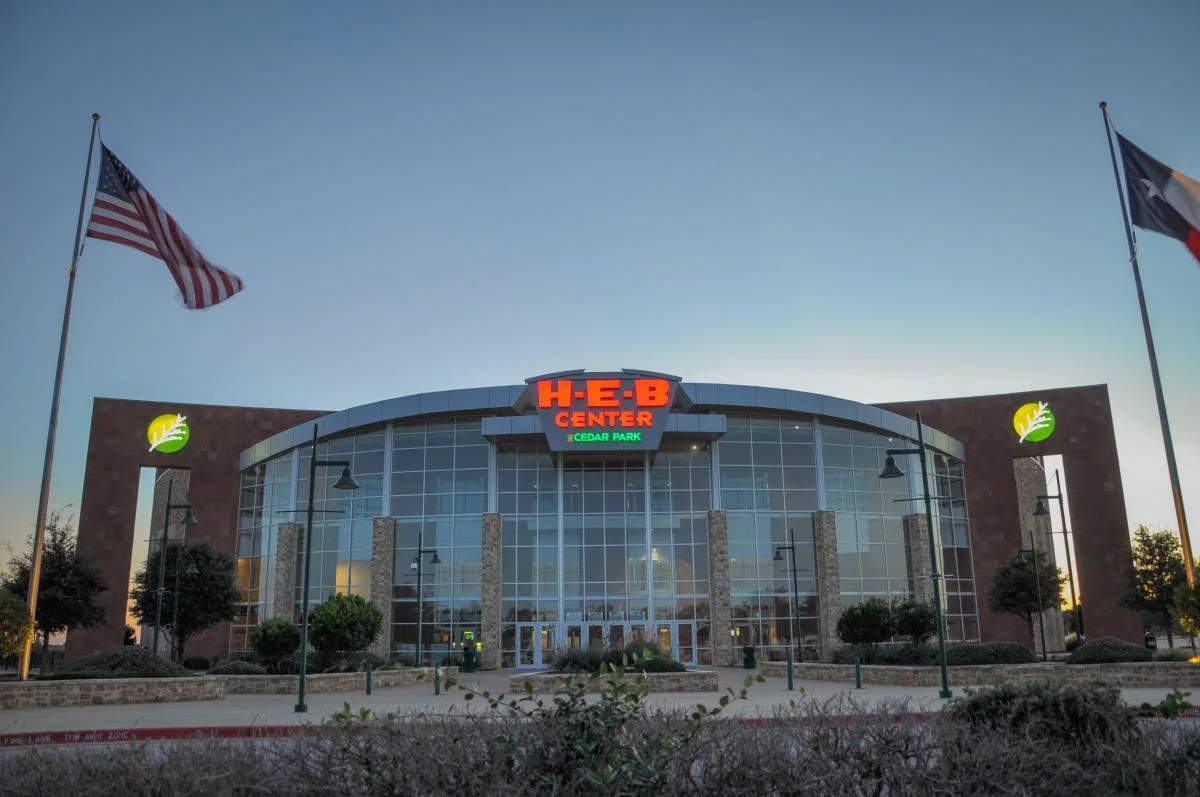 Front view of a modern H-E-B grocery store with glass facade, American and Texas flags flying nearby, and the store sign illuminated at dusk.