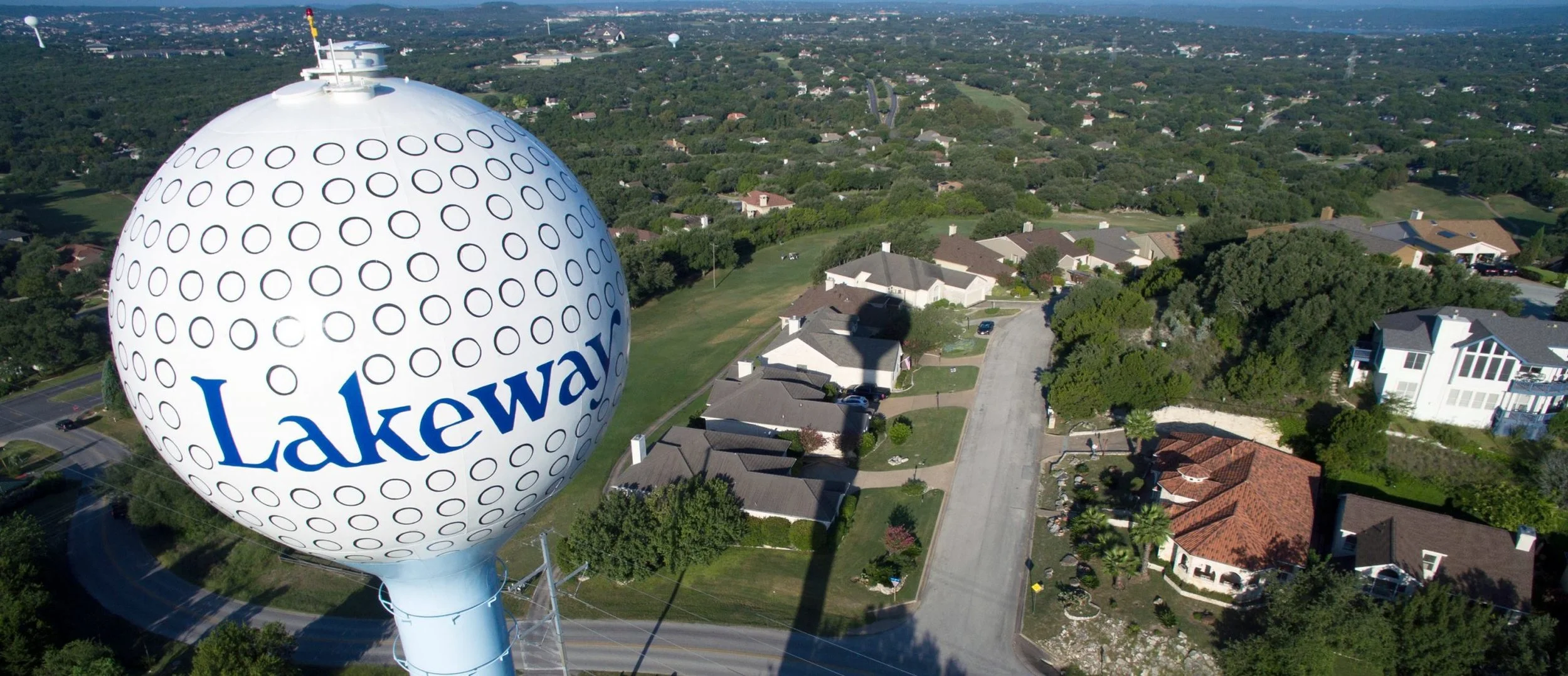 A large water tower with a spherical top, showcasing the Lakeway logo, is situated above a suburban neighborhood with houses, trees, and a curving street.