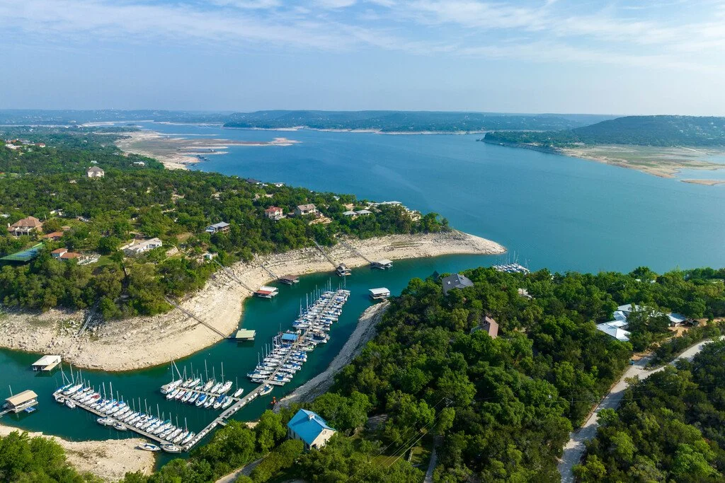 Aerial view of a marina with boats docked along a shoreline, surrounded by green trees and residential houses, overlooking a large river or lake.