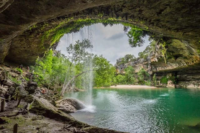 View from inside a cave looking out at a river surrounded by green trees and a rocky landscape.