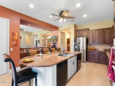 Open kitchen with granite countertop island, wooden cabinets, stainless steel refrigerator, and dining area with chairs and a ceiling fan.