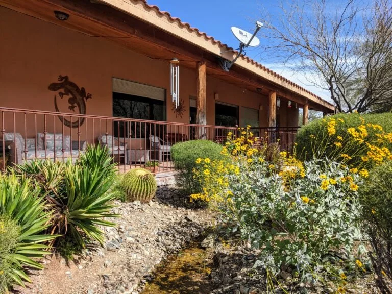 Front view of a house with a patio, decorated with Western-style wall hanging, called wind chime, and a satellite dish on the roof. The yard features desert plants, including a cactus, yucca, and yellow blooming shrubs, with rocks and gravel ground.