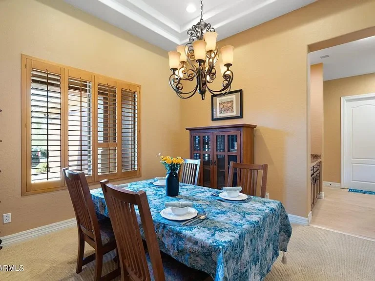 Dining room with a wooden table covered by a blue floral tablecloth, decorated with a blue vase of yellow flowers, surrounded by four wooden chairs. There is a wooden cabinet against the beige wall and a black chandelier hanging from the ceiling. A window with wooden shutters is on the left side, and a doorway leading to the kitchen is on the right.