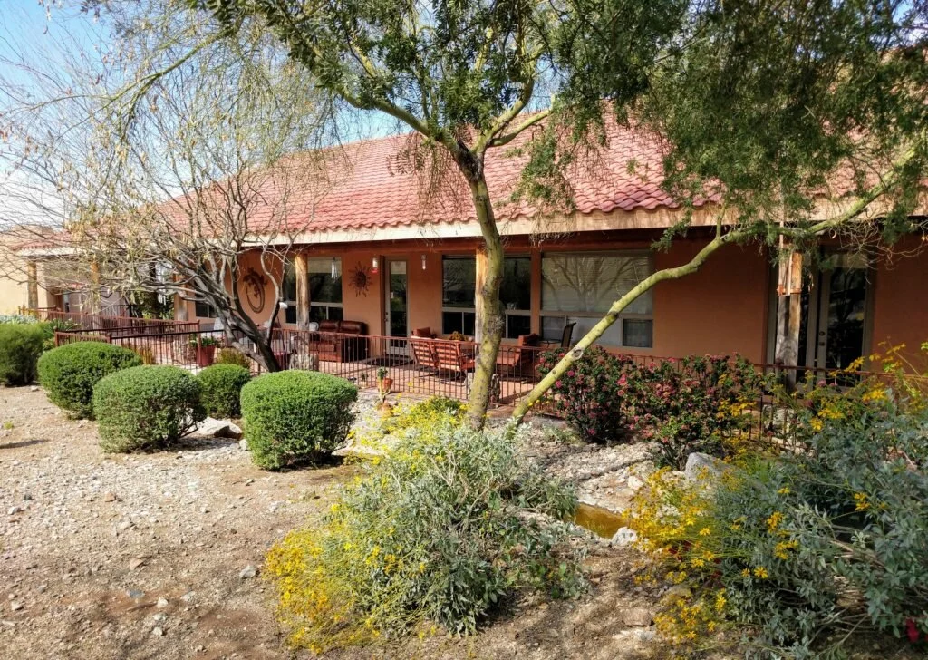 A house with a red-tiled roof, large patio with outdoor furniture, surrounded by bushes and desert plants, with trees and a clear blue sky nearby.
