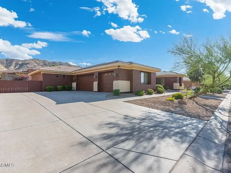 A row of single-story houses with attached garages, a concrete driveway, desert landscaping, a large tree, and a mountain range in the background under a partly cloudy blue sky.