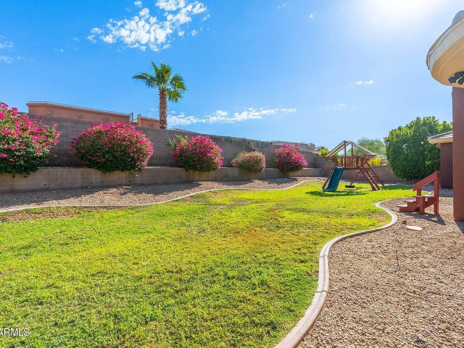 A backyard with a green lawn, colorful flowering bushes, a small play structure with a slide, and a gravel pathway under a blue sky with a few clouds.