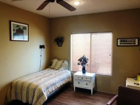 A bedroom with a single bed, a window with blinds, a white nightstand with a vase of flowers, a picture hanging on the wall, a ceiling fan, and a ceiling light.
