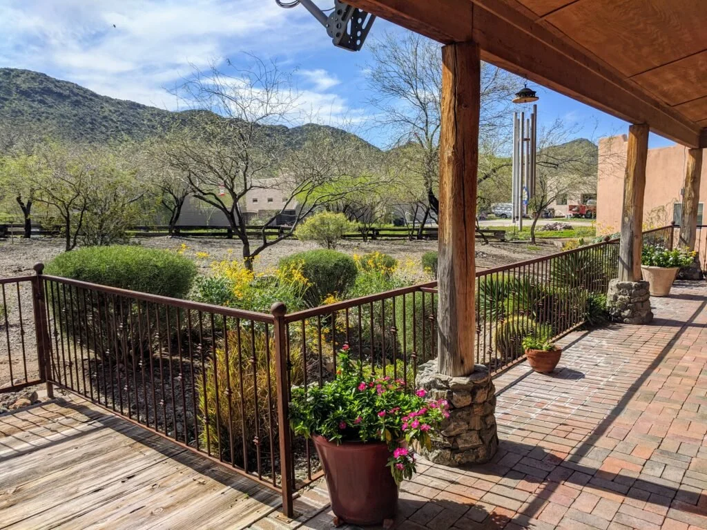 View from a porch overlooking a garden and mountains in the background, with trees and yellow flowers, under a partly cloudy sky.