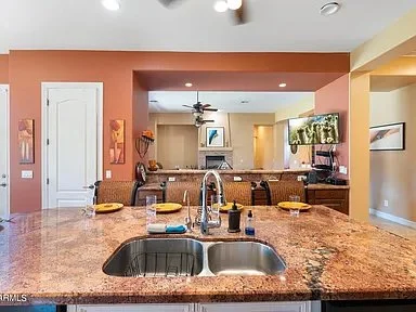 Kitchen with granite island, double sink, and place settings with yellow napkins, overlooking a living area with a wall-mounted TV and artwork.