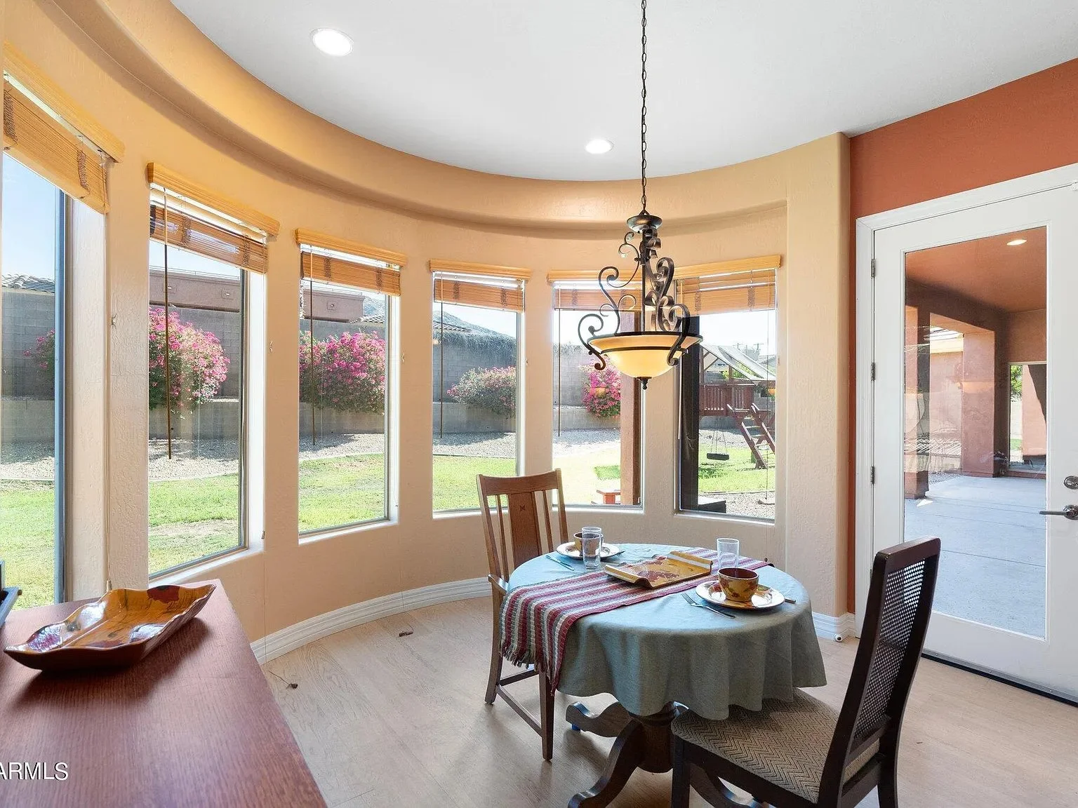 Sunlit dining area with a round table, four chairs, and a view of a backyard with flowering bushes through a series of windows.