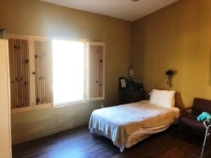 A simple bedroom with a single bed, a wooden nightstand, and a small sofa. There is a window with wooden shutters allowing light into the room.