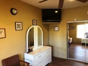 Interior of a room with a white dresser and mirror, a wall clock, framed pictures, a television mounted on the wall, and a big mirror reflecting part of the room and an adjacent room with windows and blinds.